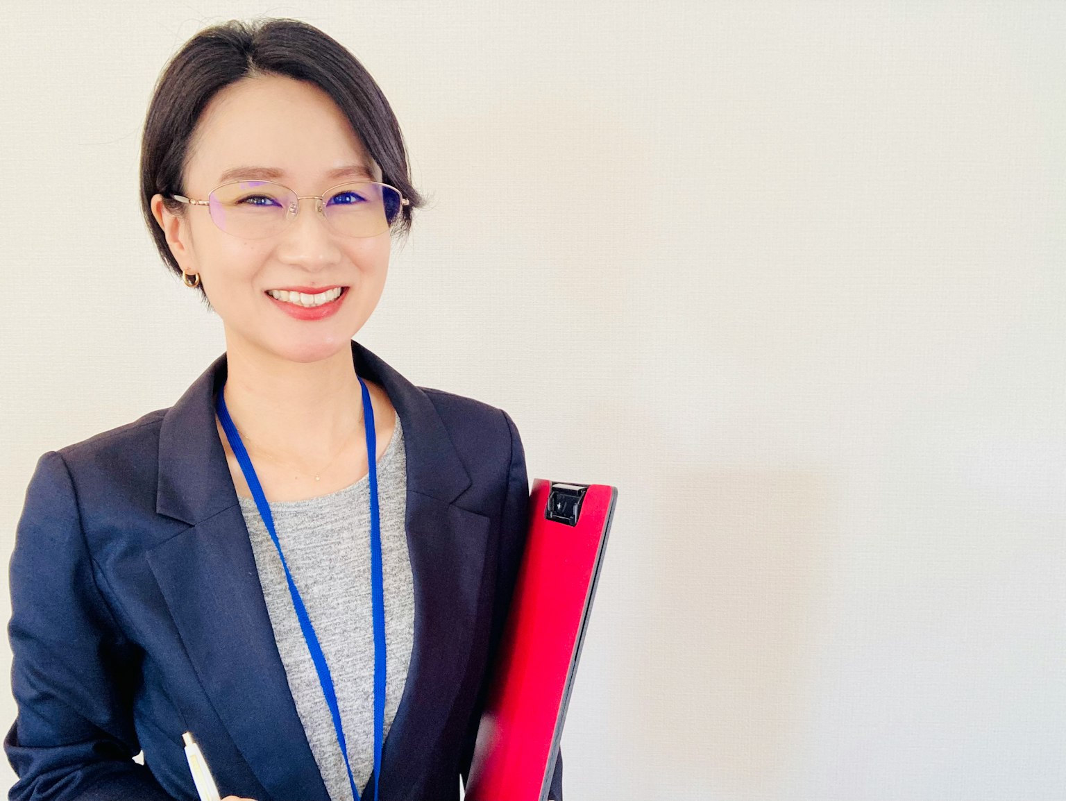 一位穿着西装的微笑女性手持红色文件夹，站在简单背景前