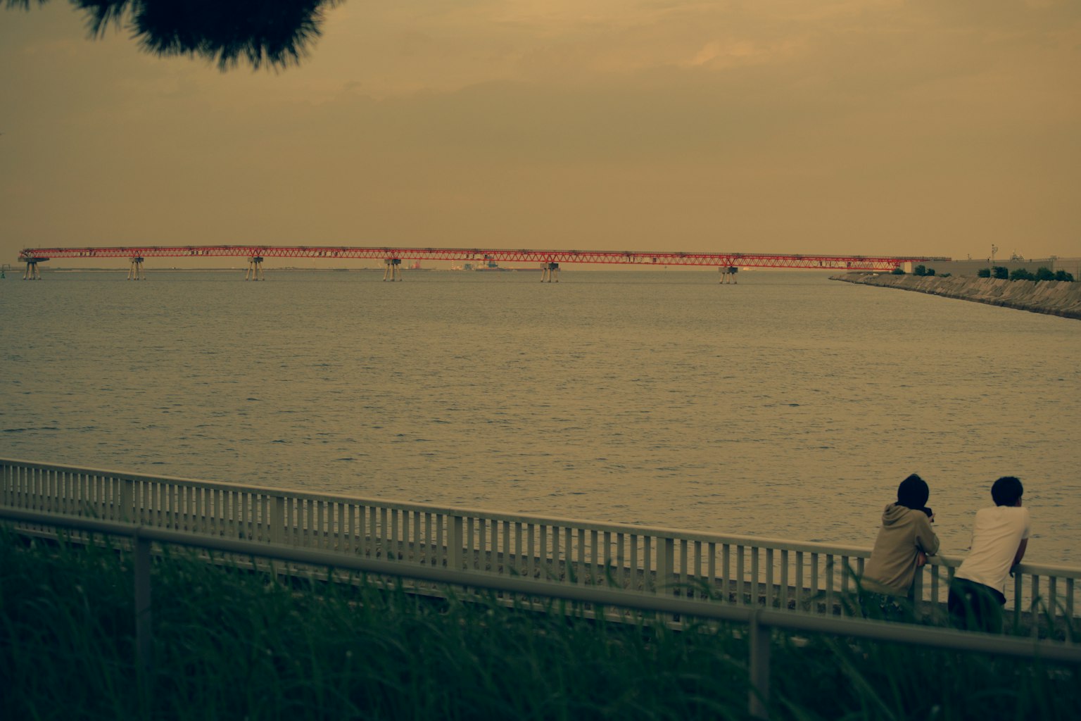 Two people sitting by the river with a red bridge in the background