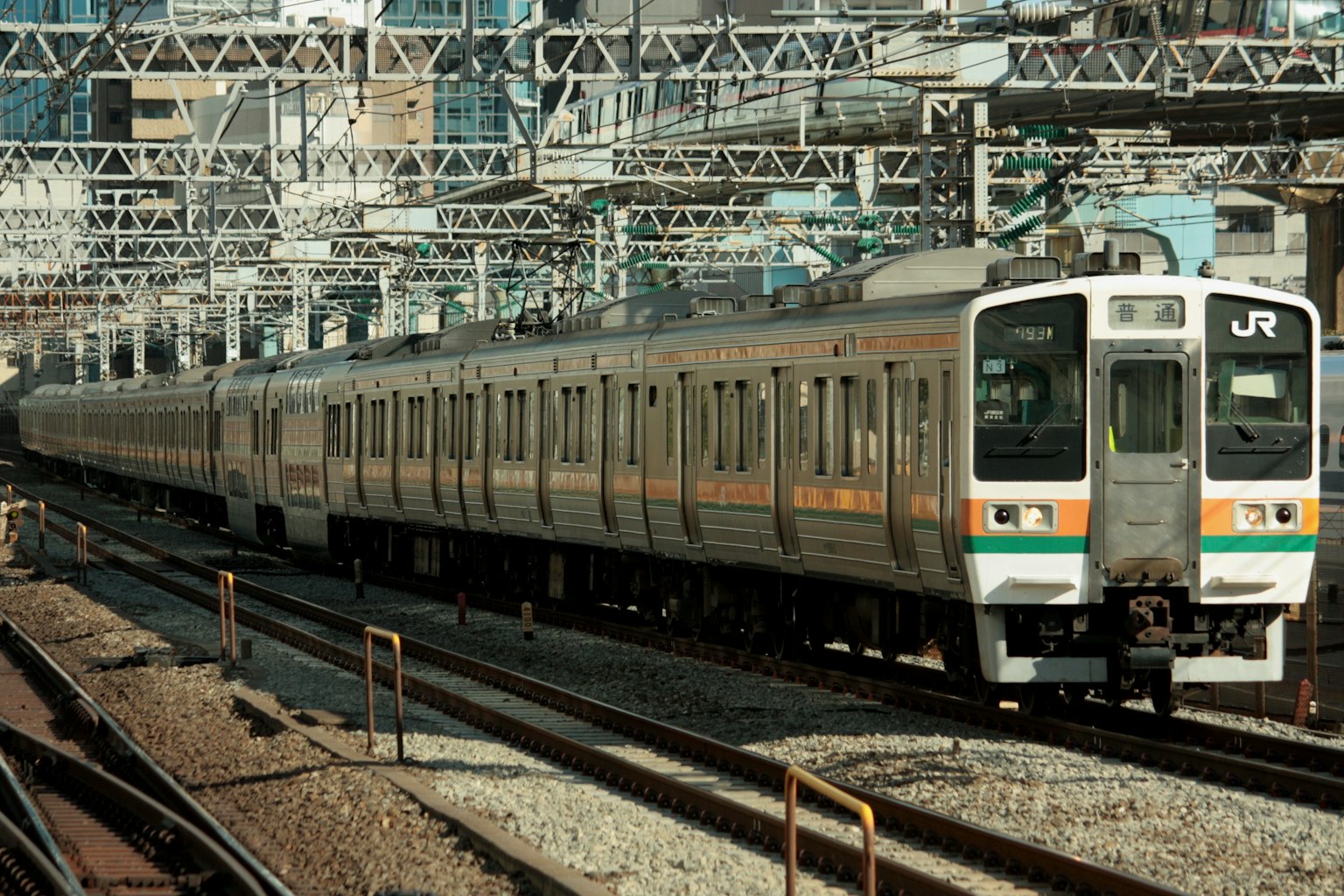 รถไฟ JR ญี่ปุ่นที่วิ่งบนรางพร้อมโครงสร้างรถไฟเหนือศีรษะในพื้นหลัง
