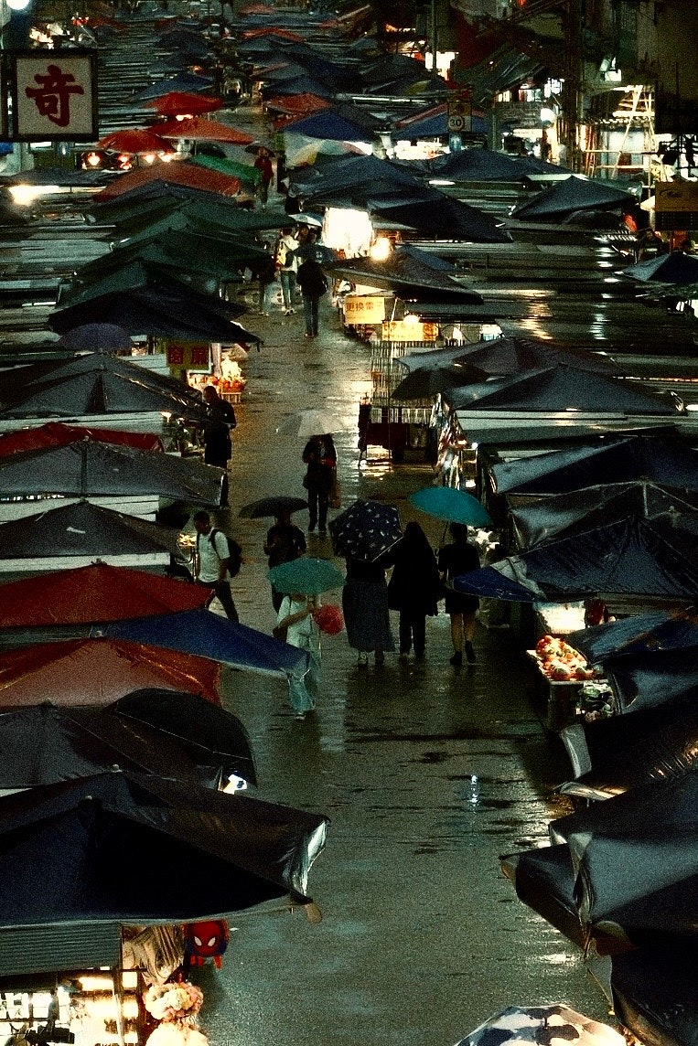 雨に濡れた市場の通りに傘を持った人々が歩いている