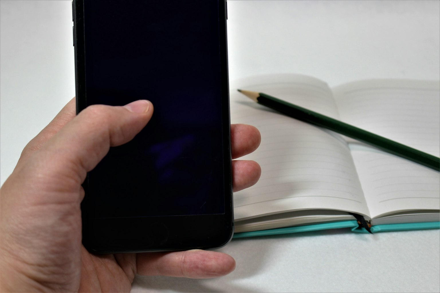 Bild einer Hand, die ein Smartphone hält, mit einem offenen Notizbuch und einem grünen Bleistift