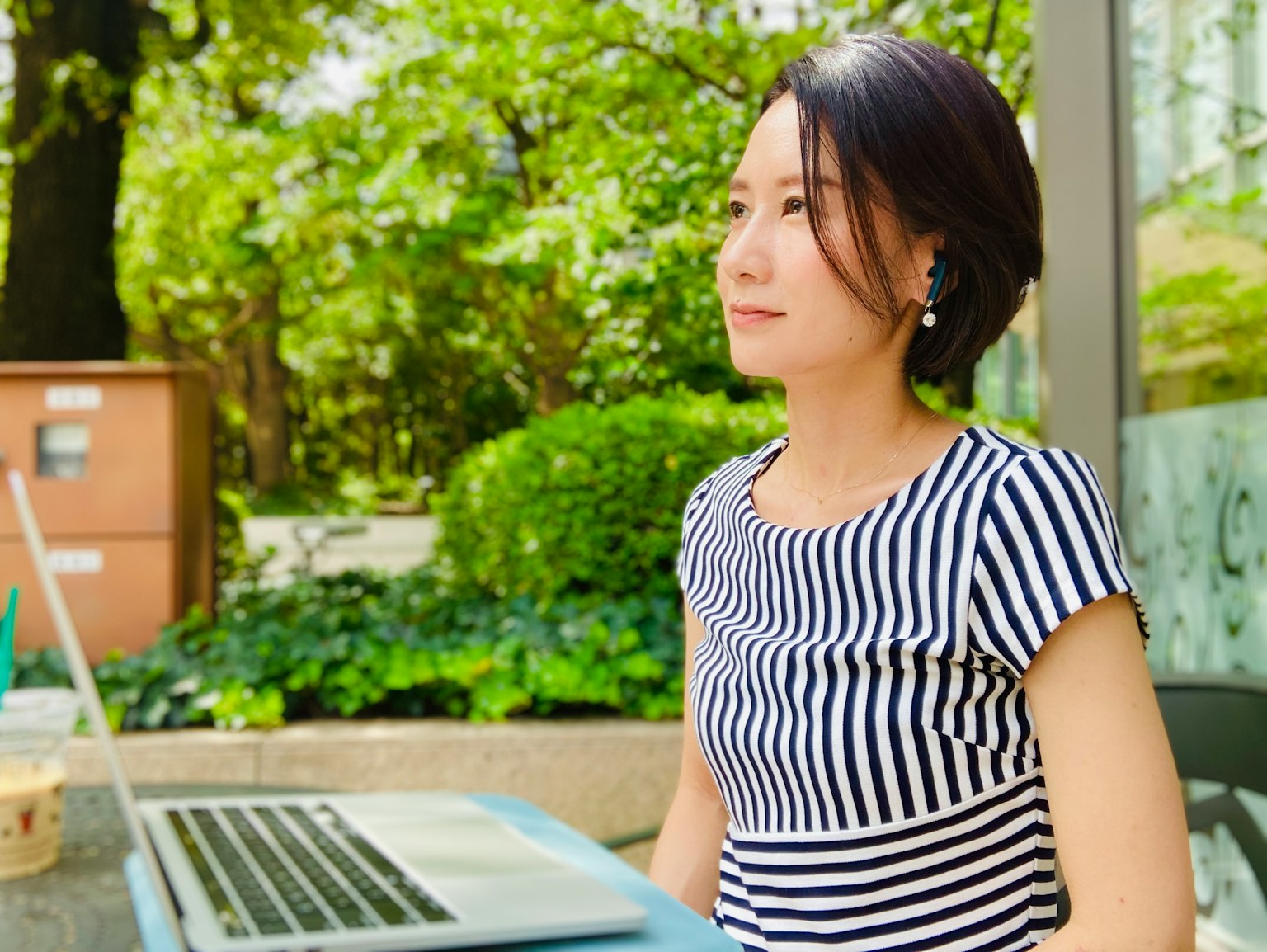 Frau, die auf einer Terrasse in einem Park einen Laptop benutzt, trägt ein gestreiftes Oberteil, umgeben von grünen Pflanzen
