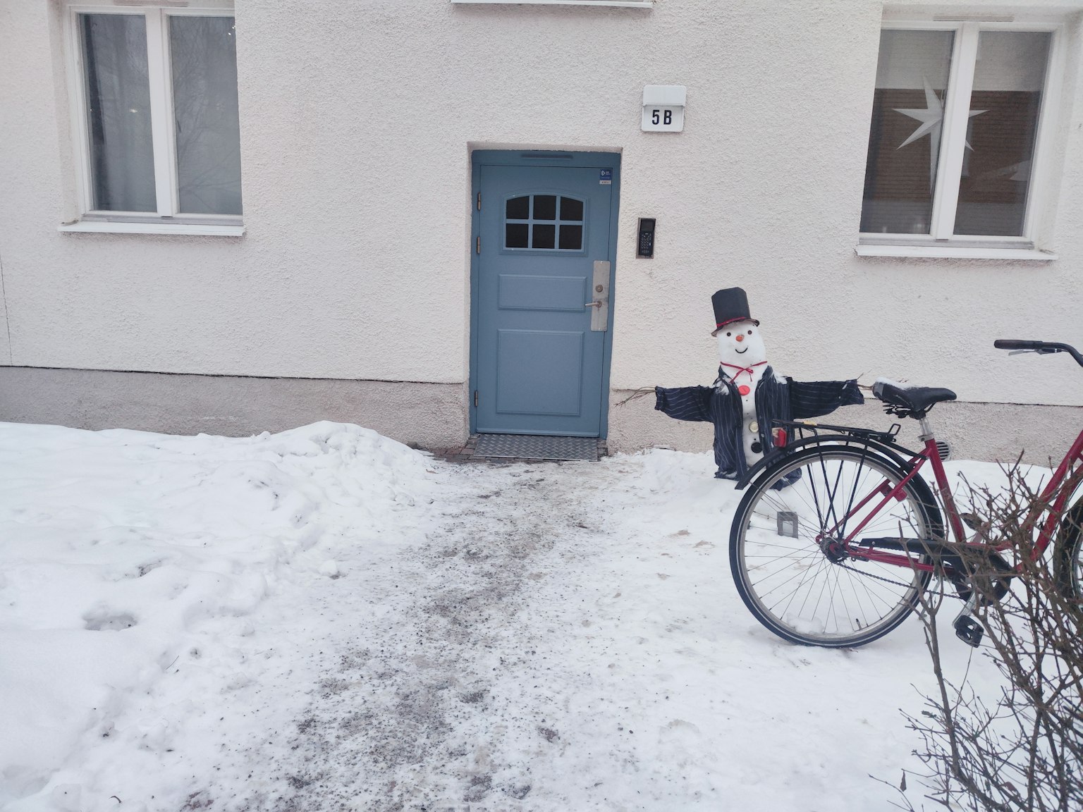 Un bonhomme de neige et un vélo devant une porte bleue recouverte de neige