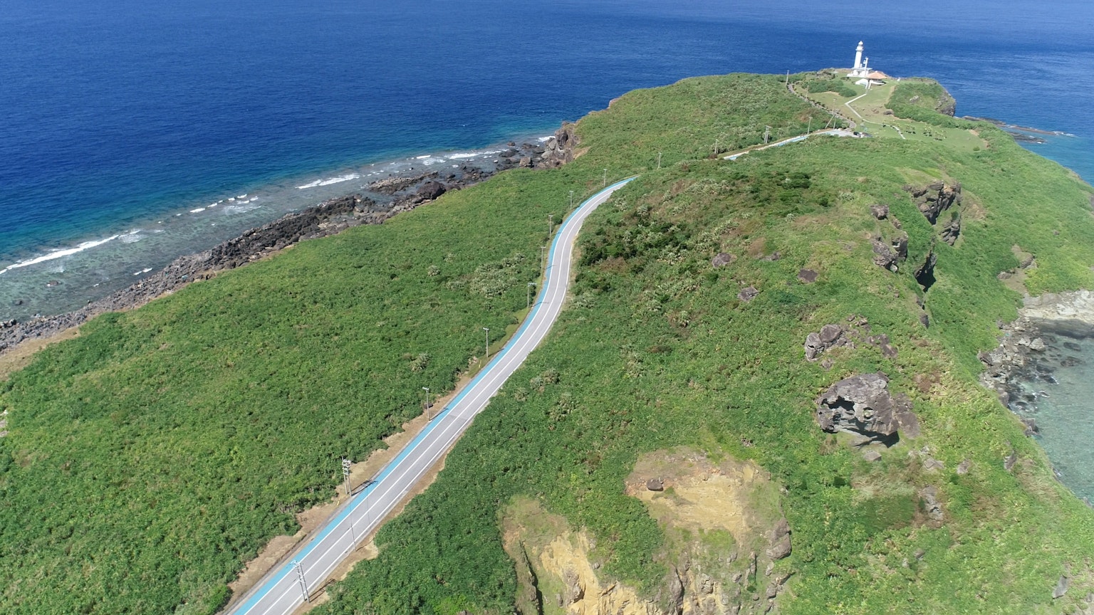 Pemandangan udara jalan berliku di bukit hijau dengan mercusuar yang menghadap ke laut biru
