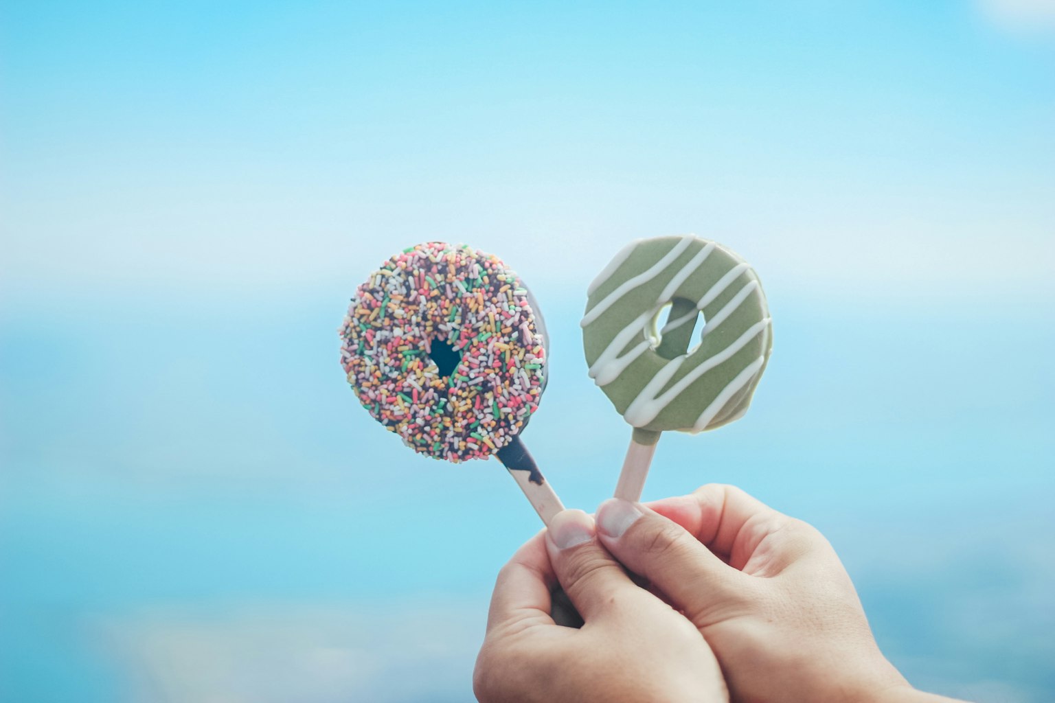 Zwei bunte Donut-förmige Eis am Stiel, die von Händen vor einem blauen Himmel gehalten werden
