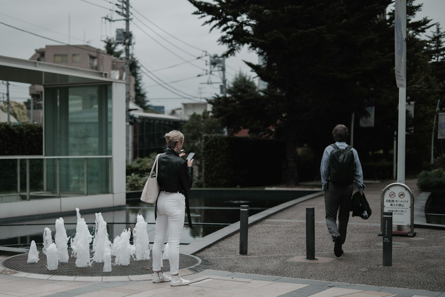 Two individuals walking on a cloudy day near a modern building with white fountains
