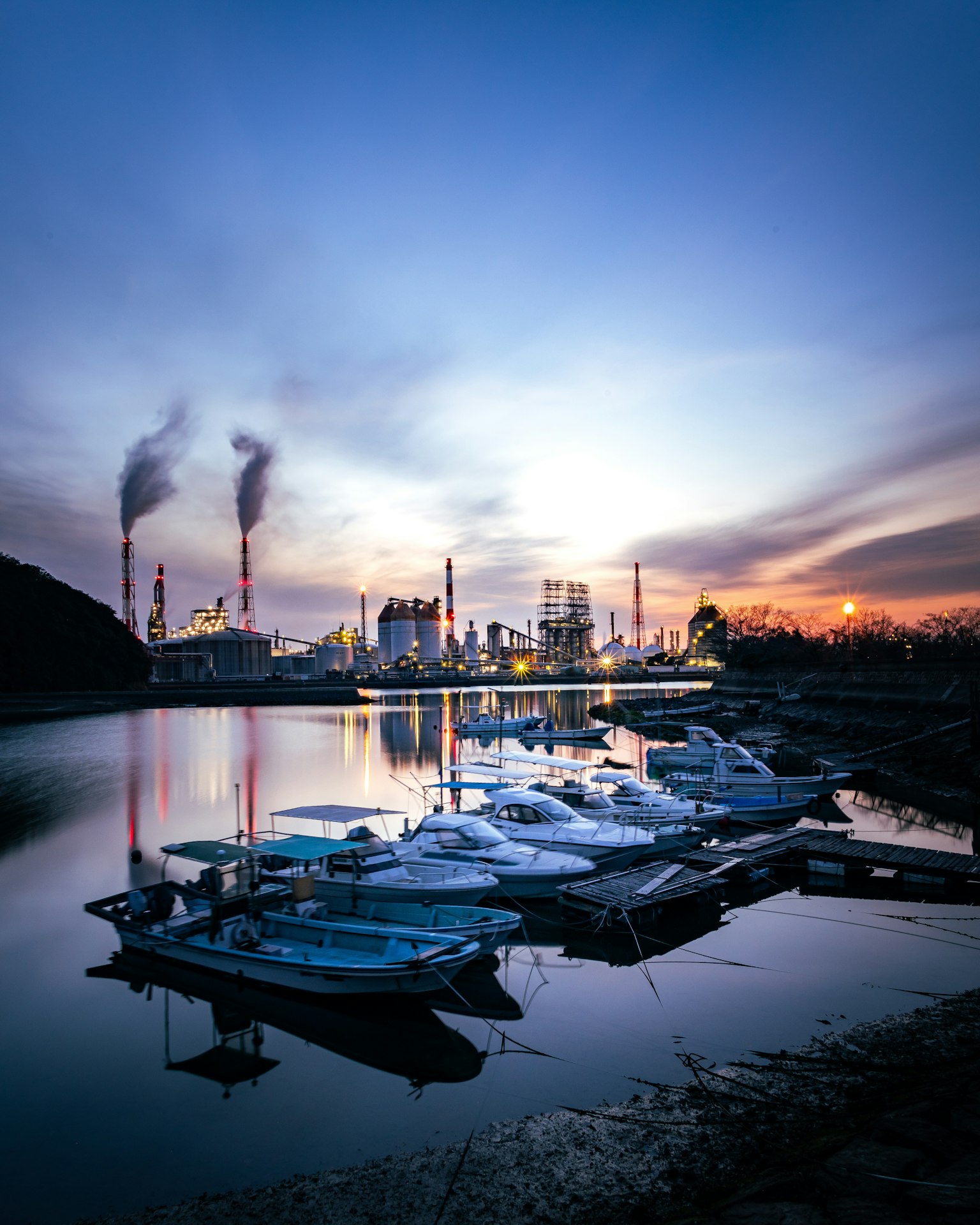 Boote am Ufer eines Flusses mit Industrie-Schornsteinen bei Sonnenuntergang