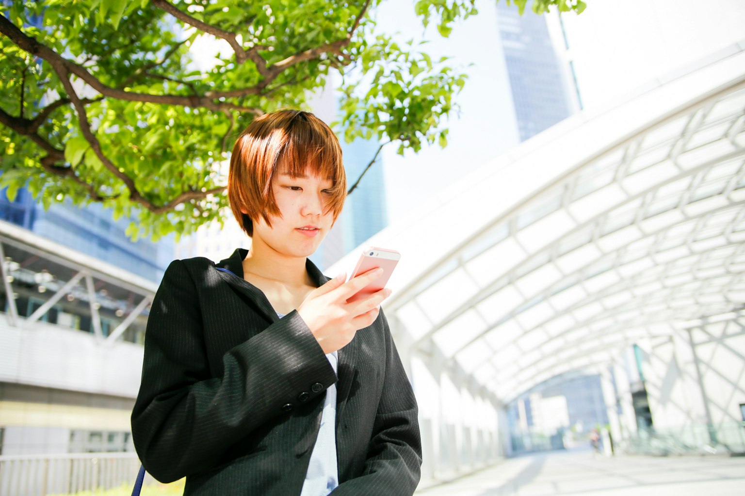 Người phụ nữ trẻ nhìn vào điện thoại dưới gốc cây xanh với kiến trúc hiện đại phía sau