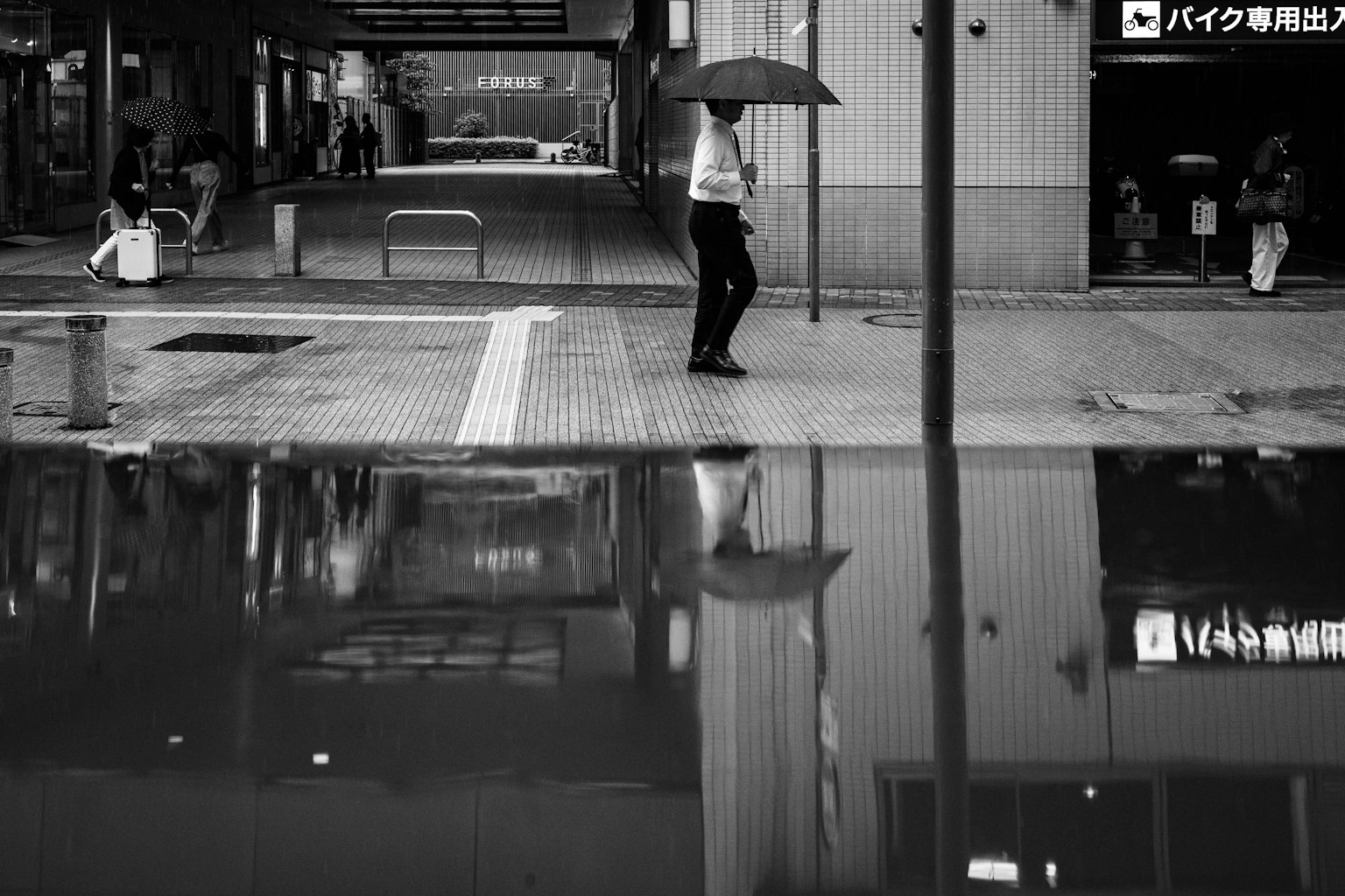 雨の中で傘を持って歩く人の白黒写真 水たまりに映る反射が印象的