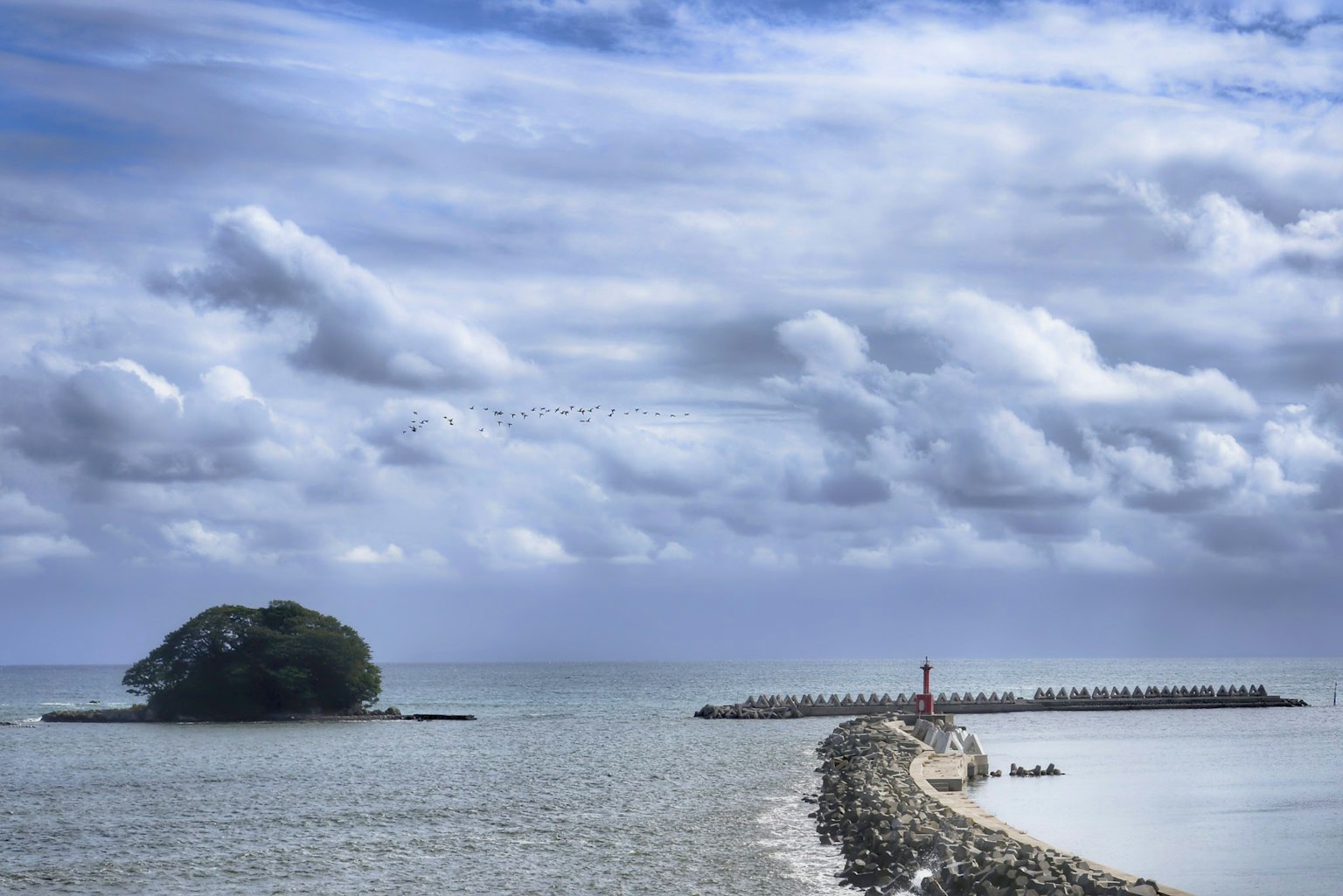 View of a small island and breakwater under a blue sky with clouds