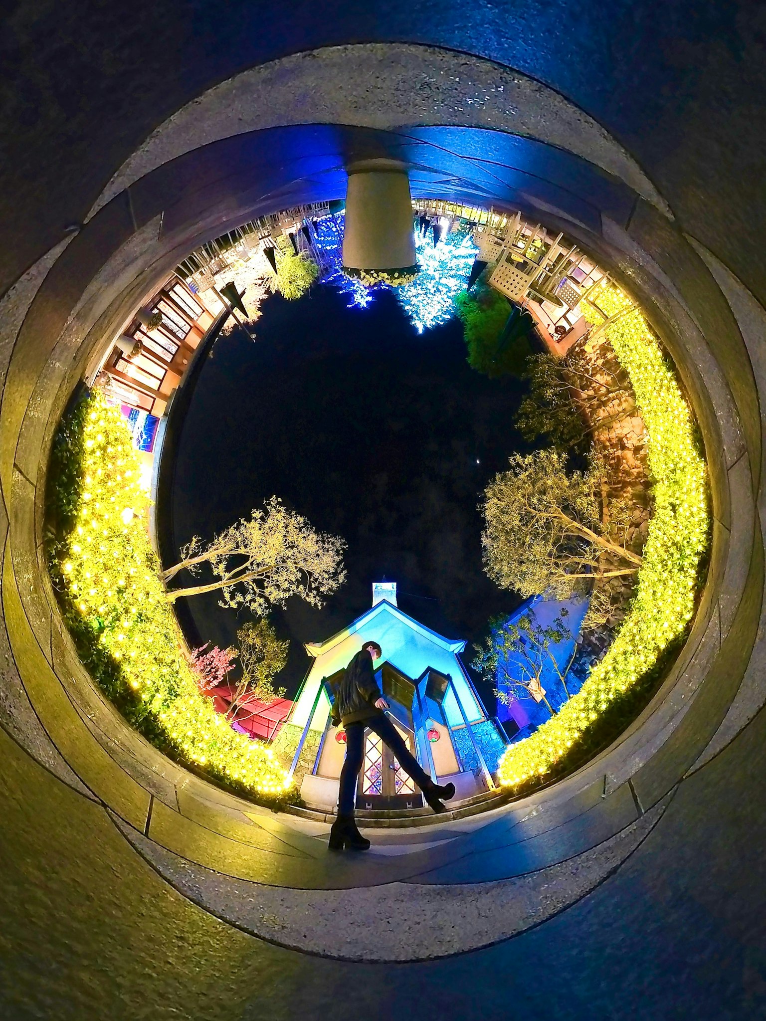 Pareja de pie en una vista circular de un parque por la noche con árboles iluminados y una pequeña cabaña