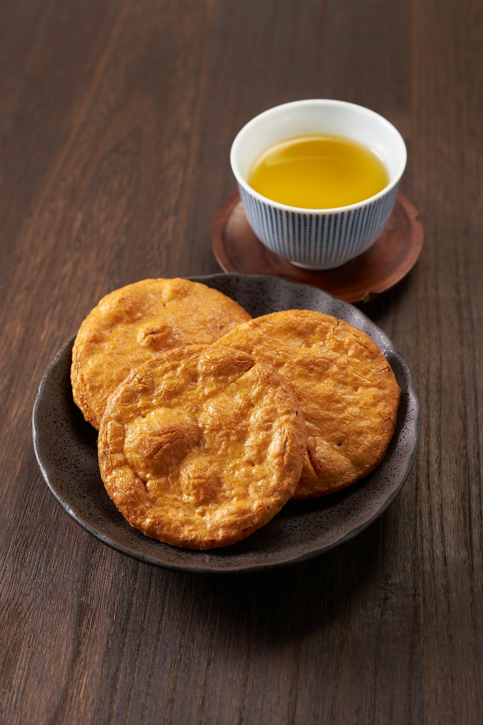Tres galletas recién horneadas en un plato oscuro junto a una taza de té verde sobre una mesa de madera