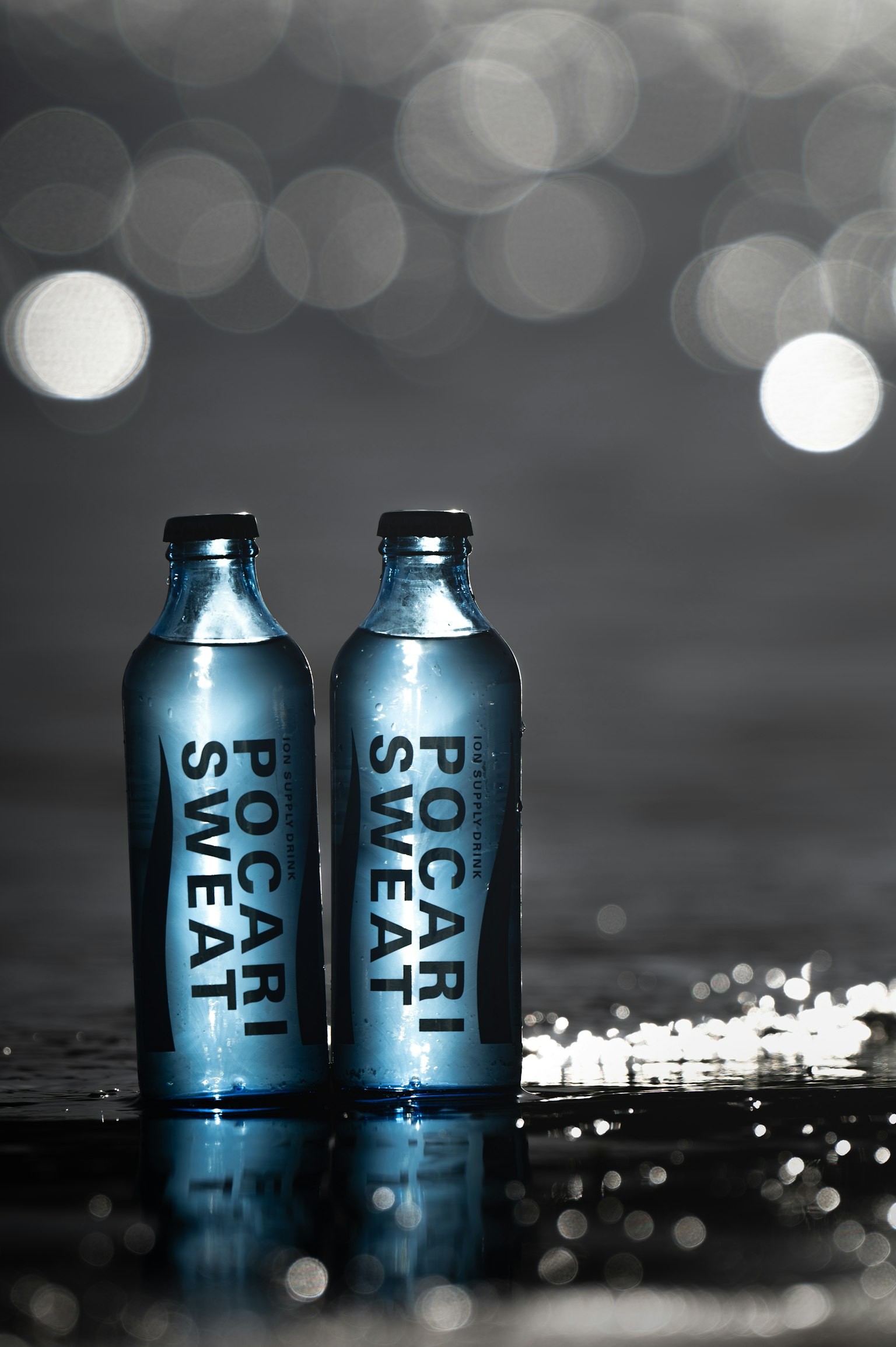 ขวดน้ำสีฟ้าสองขวดของโปคาริ สเวต บนพื้นผิวสะท้อนกับแสงเบลอในพื้นหลัง