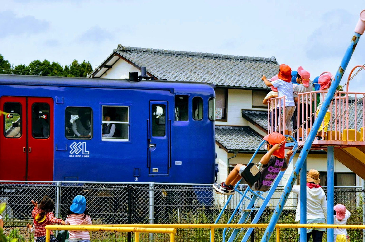 Un train bleu passant devant un terrain de jeu avec des enfants jouant
