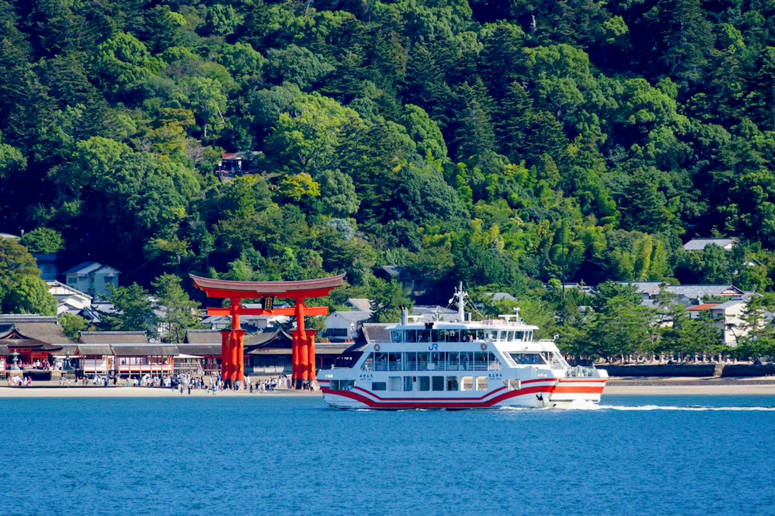 Un ferry navegando en aguas azules cerca de montañas verdes y una puerta torii roja