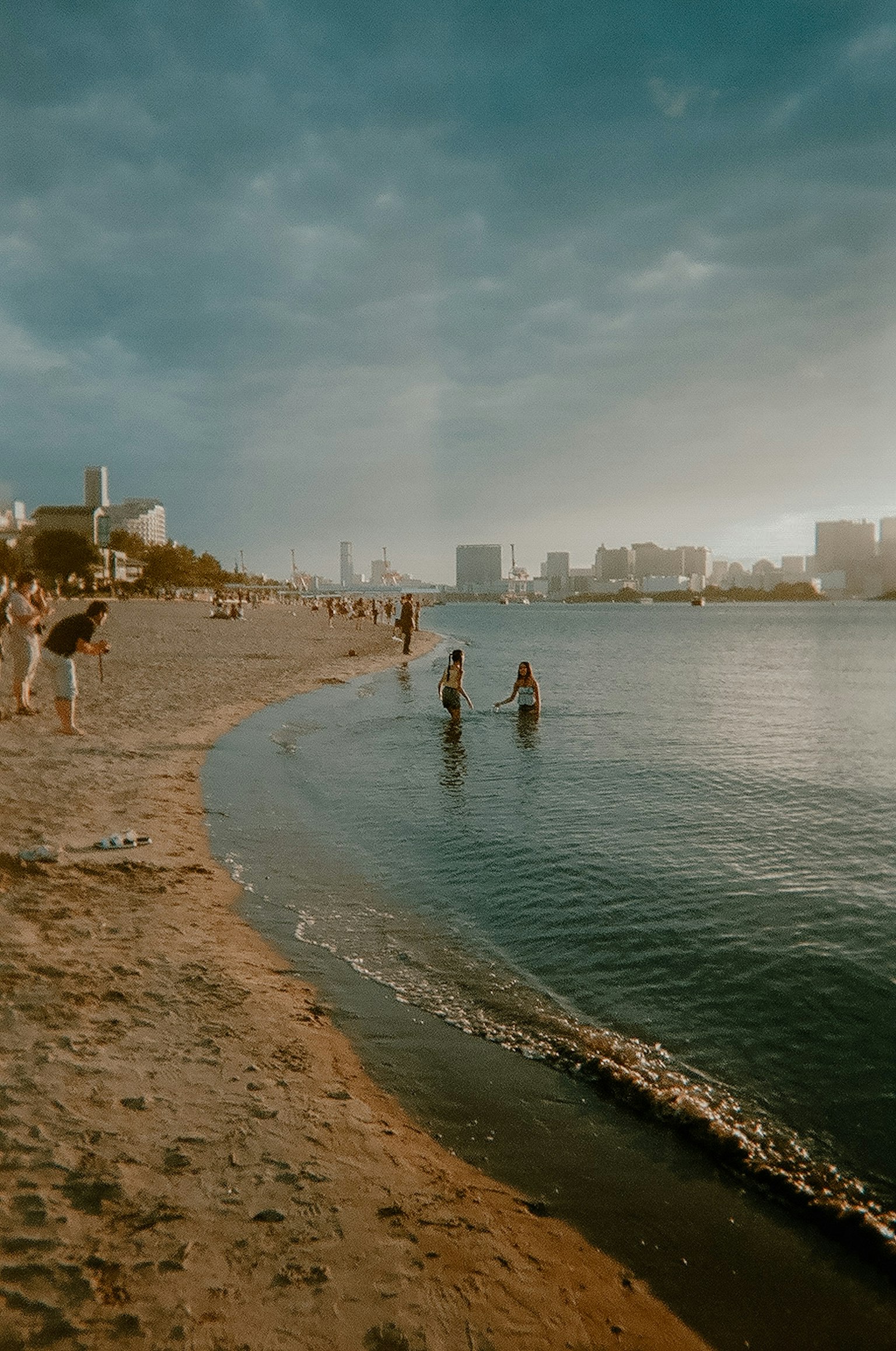 Personas disfrutando de la playa con agua tranquila y un horizonte urbano a lo lejos