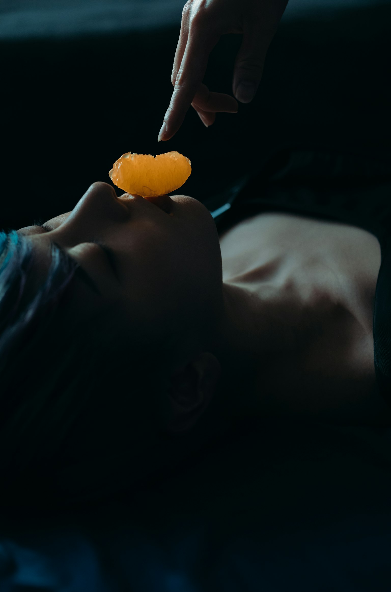 A woman lying against a dark background with an orange slice on her lips