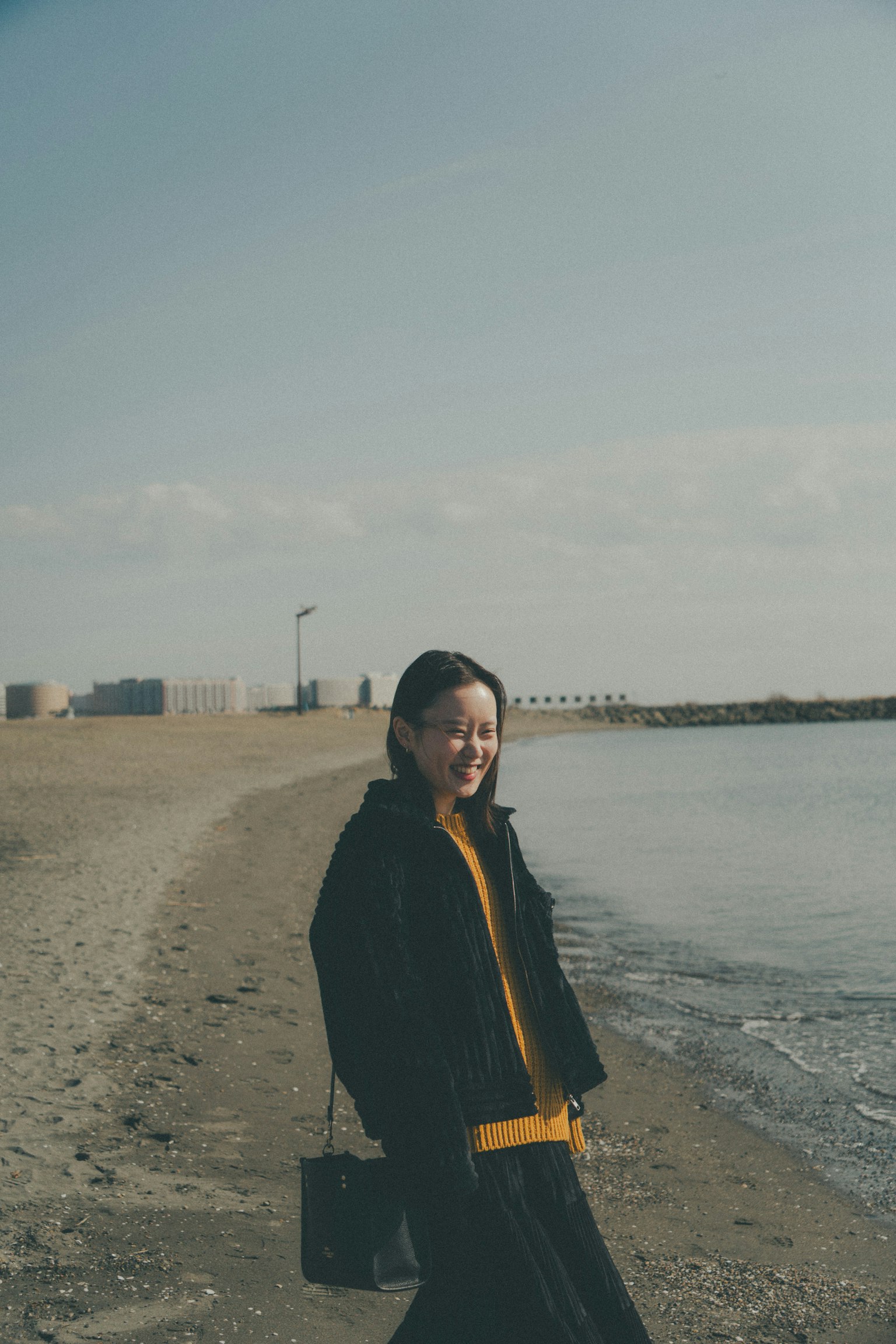 Una donna sorridente sulla spiaggia che indossa una giacca nera e un maglione giallo