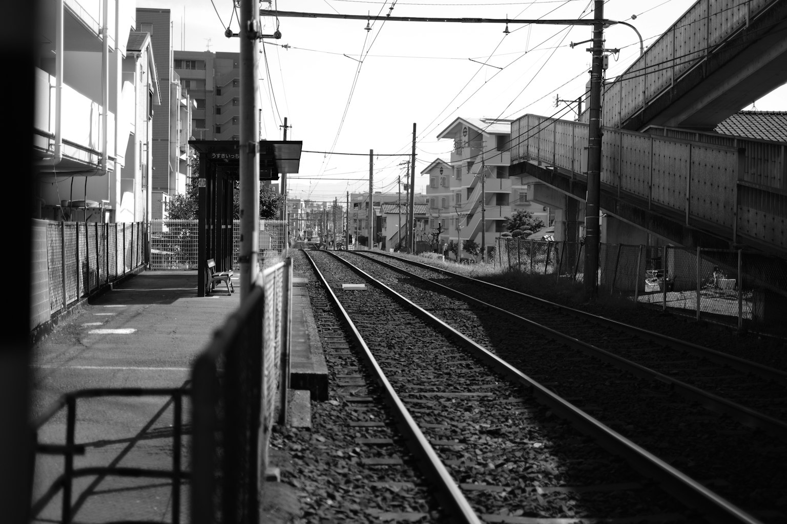 Scène ferroviaire en noir et blanc avec des voies ferrées et un quai de gare bâtiments à proximité et un viaduc