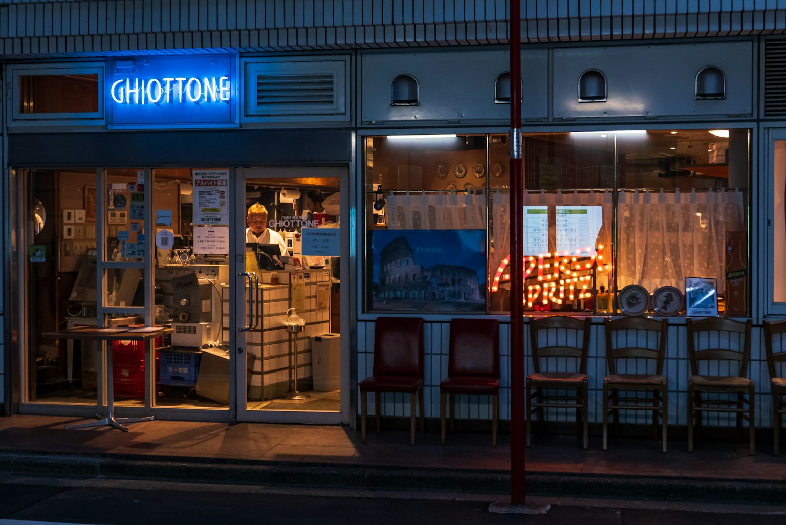 Ngoại thất của nhà hàng GHIOTTONG vào ban đêm cửa sổ được chiếu sáng và ghế được sắp xếp quanh bàn