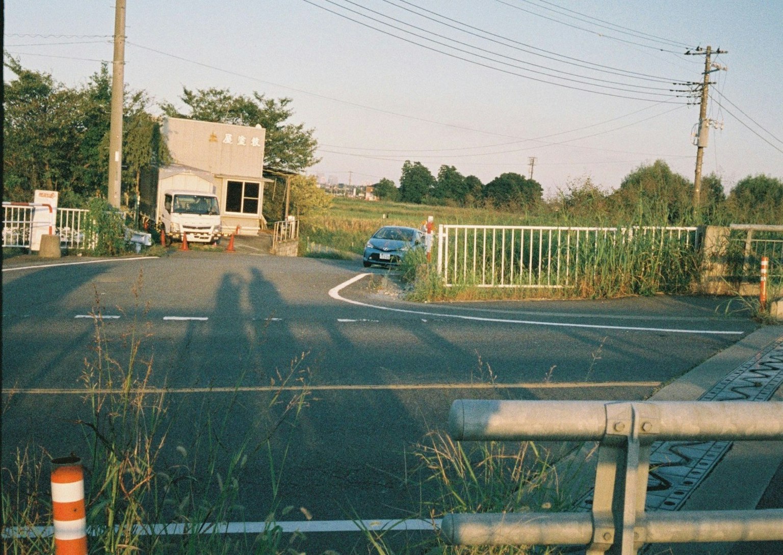 A blue car approaching an intersection with a truck and a fence in the background