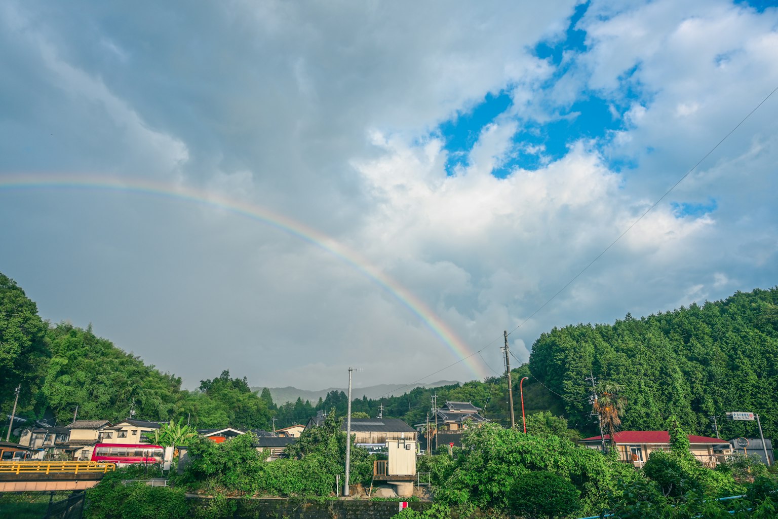 彩虹在藍天和雲朵之間拱起,綠山和房屋在其下方