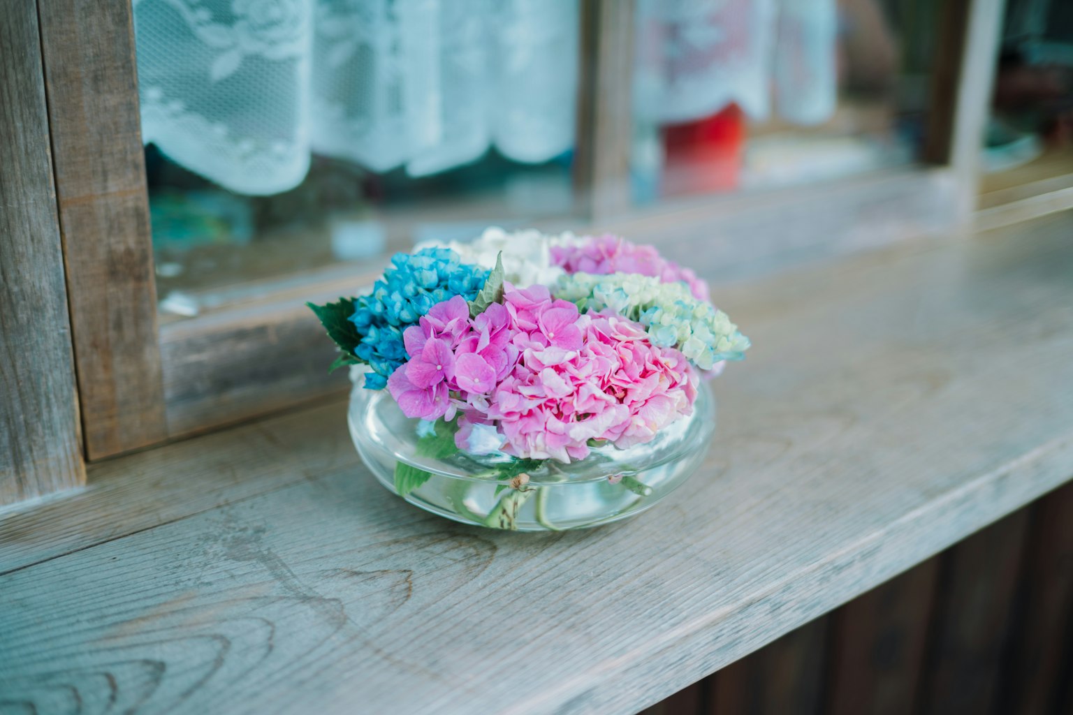 Flores coloridas dispuestas en un tazón transparente sobre una superficie de madera cerca de una ventana
