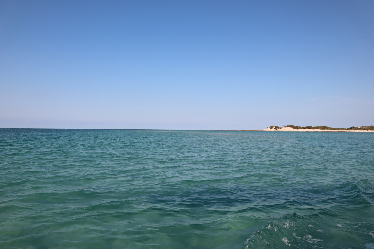 Pemandangan luas laut dan langit biru dengan gelombang lembut dan pantai berpasir di kejauhan