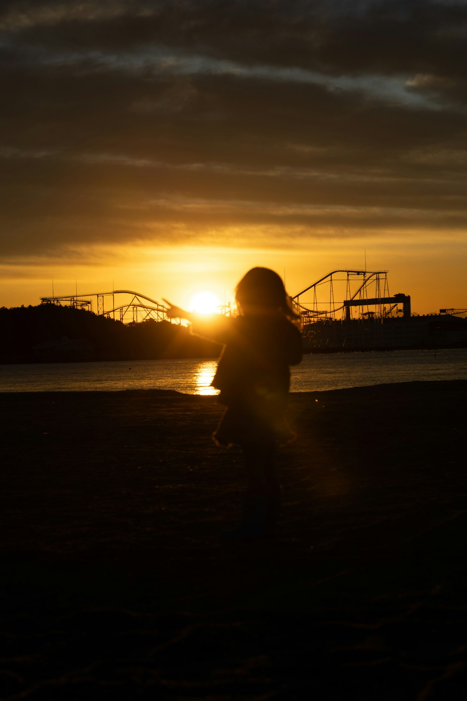 Silhouette anak melawan matahari terbenam dengan roller coaster taman hiburan di latar belakang