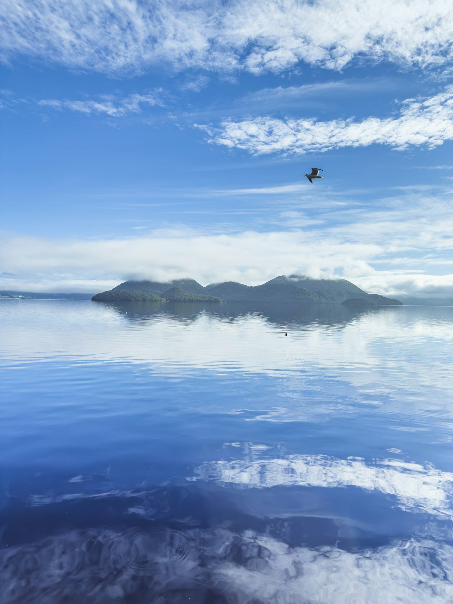 Pemandangan danau tenang yang memantulkan langit biru dan awan dengan sebuah pulau di tengah