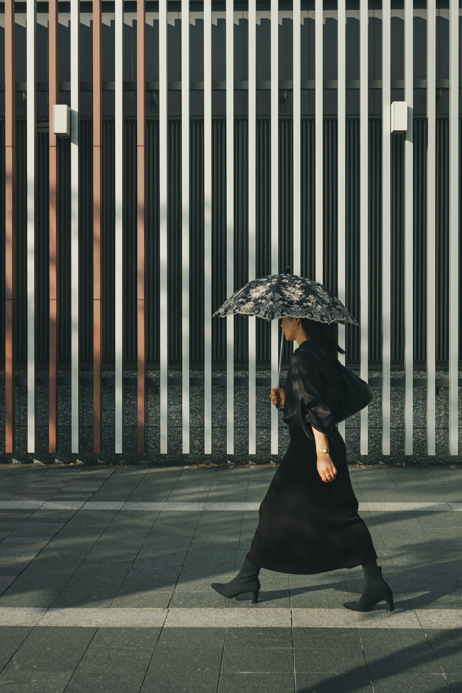 Une femme en vêtements noirs marchant avec un parapluie devant un mur rayé