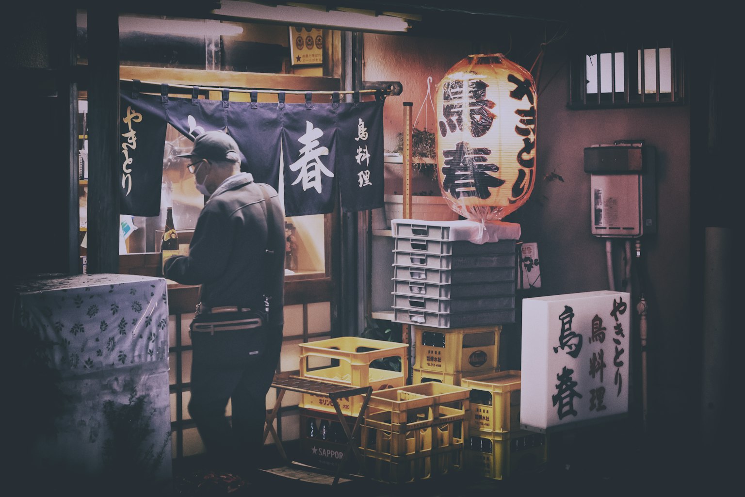Nighttime scene of a Japanese eatery man standing in front illuminated lanterns and signs