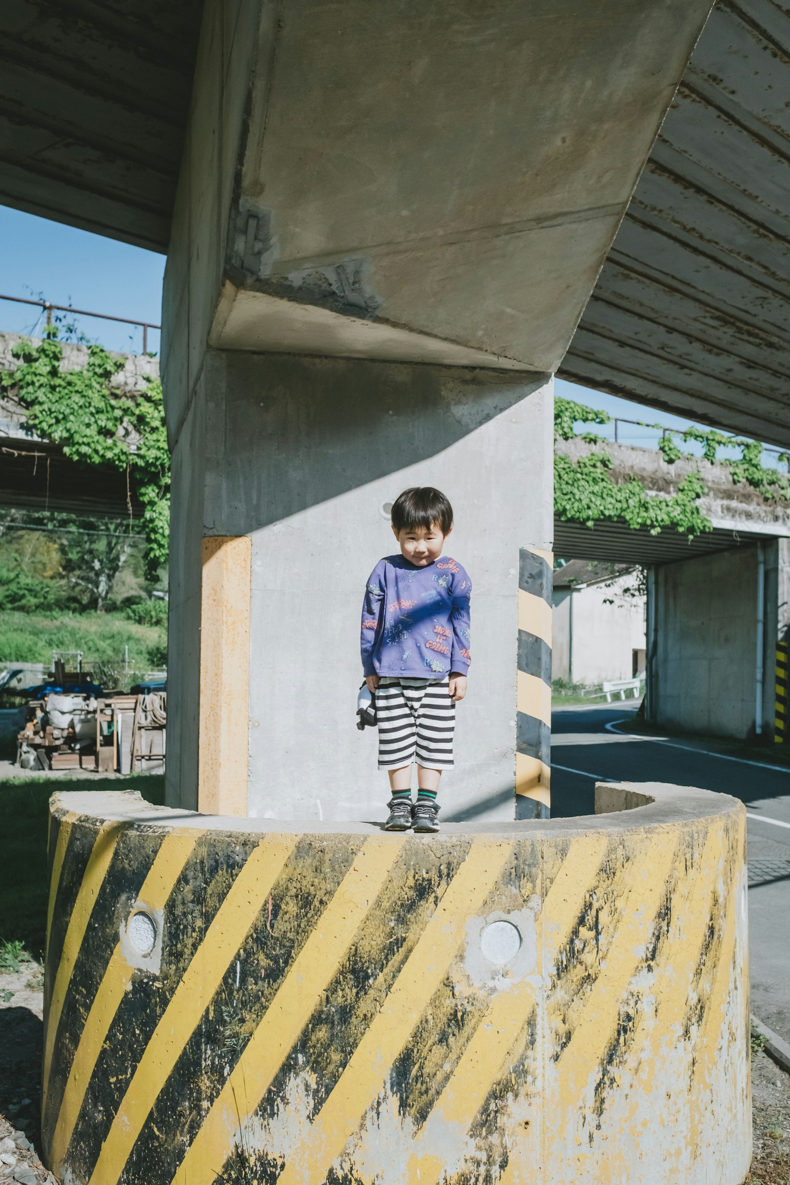 一個孩子站在一根黃色和黑色條紋的柱子上，背景是綠色的混凝土橋下