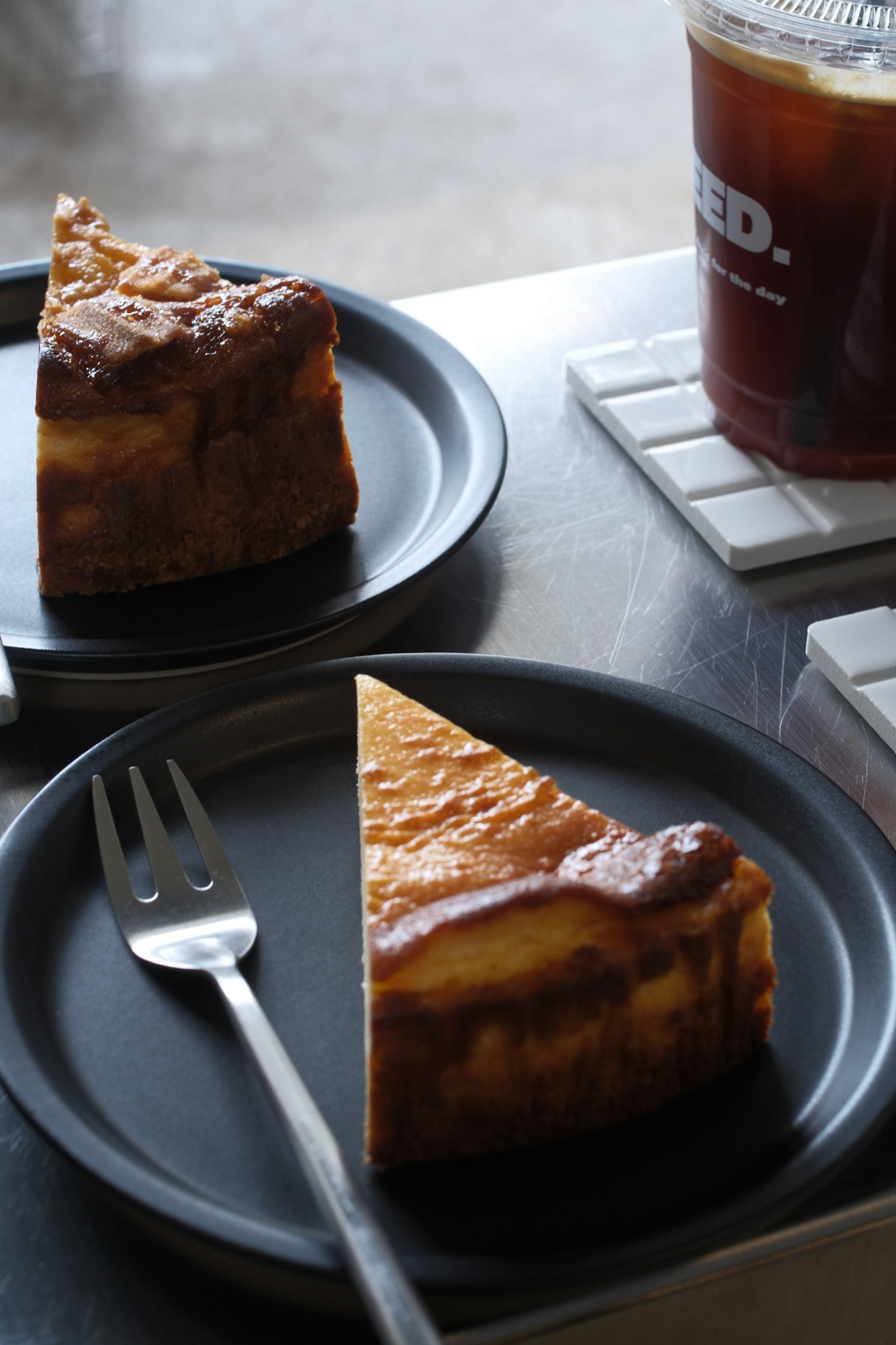 Dua potong cheesecake di piring hitam di atas meja kafe dengan segelas kopi es di latar belakang