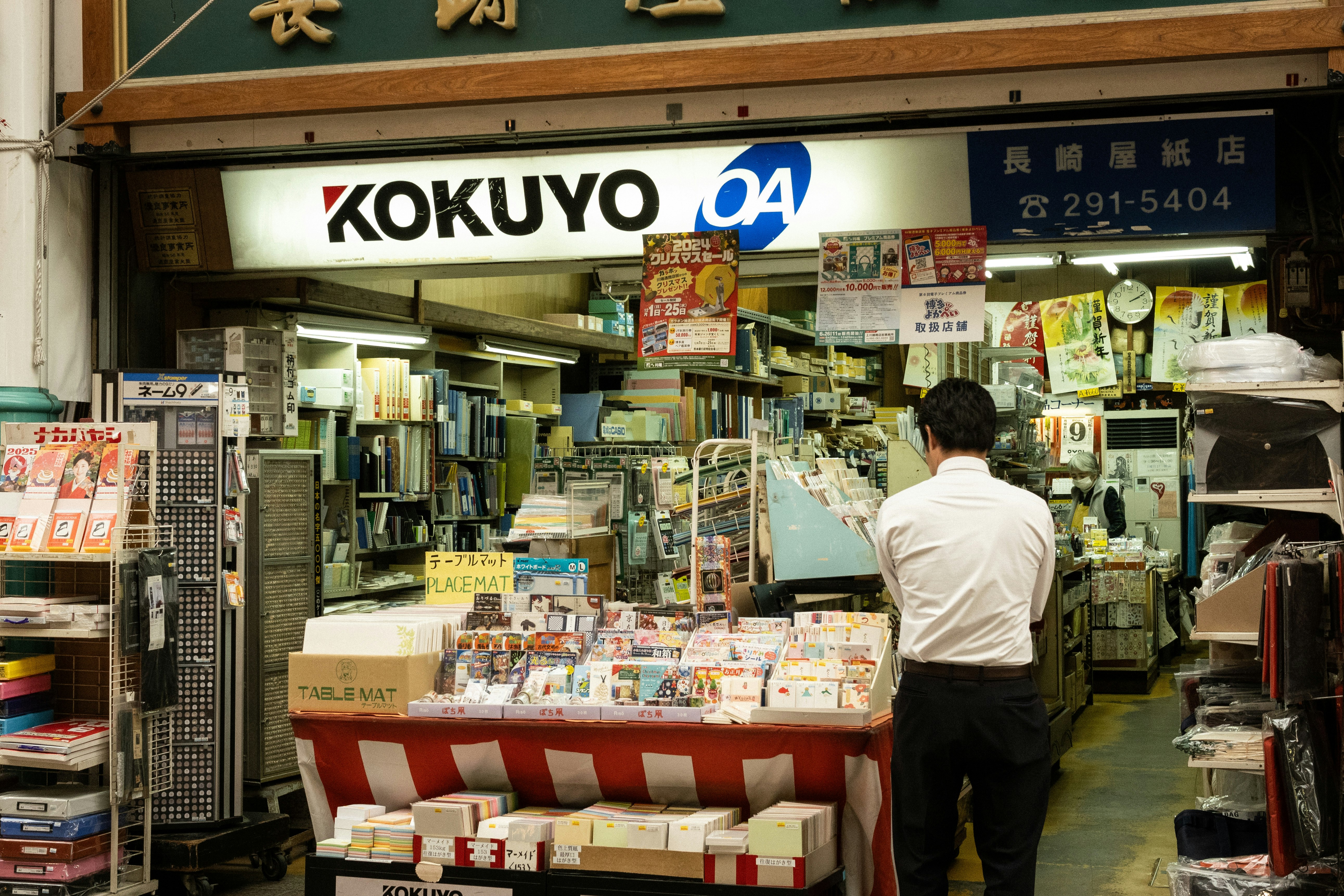 หน้าร้านร้านเครื่องเขียนพร้อมชั้นวางและป้ายในตรอกแคบ