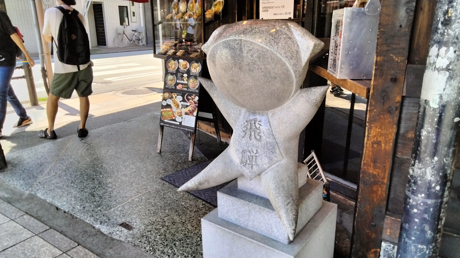 Unique sculpture standing on the sidewalk with a large circular head on a white star-shaped base pedestrians nearby