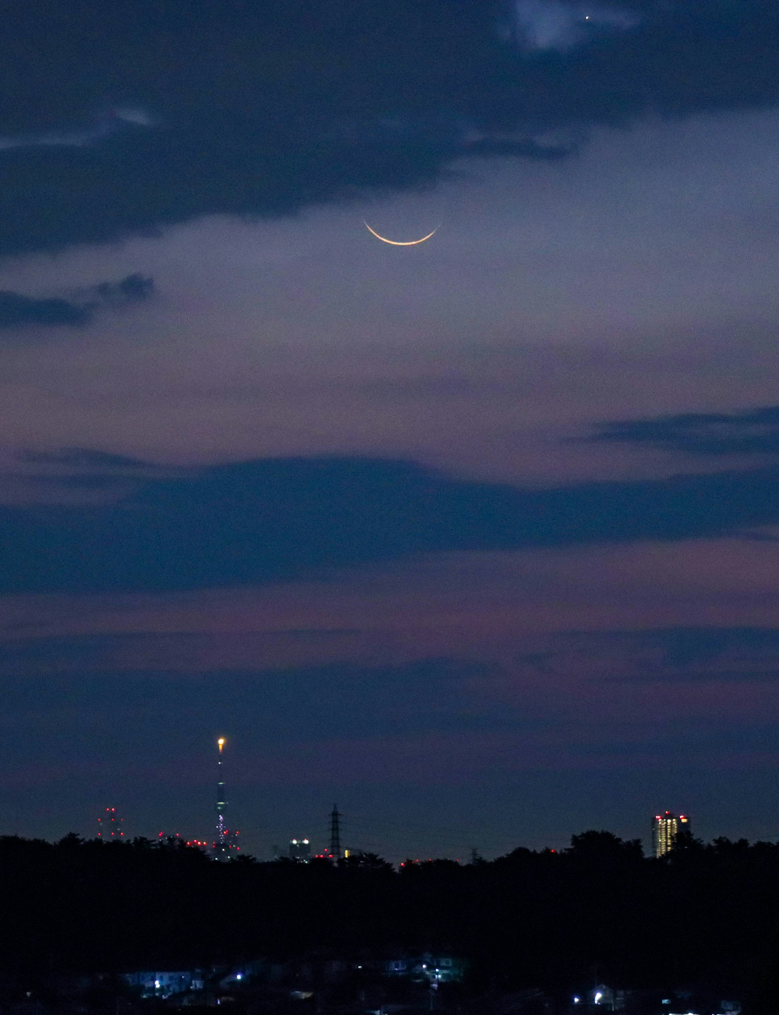 A crescent moon in a twilight sky with city silhouettes in the background