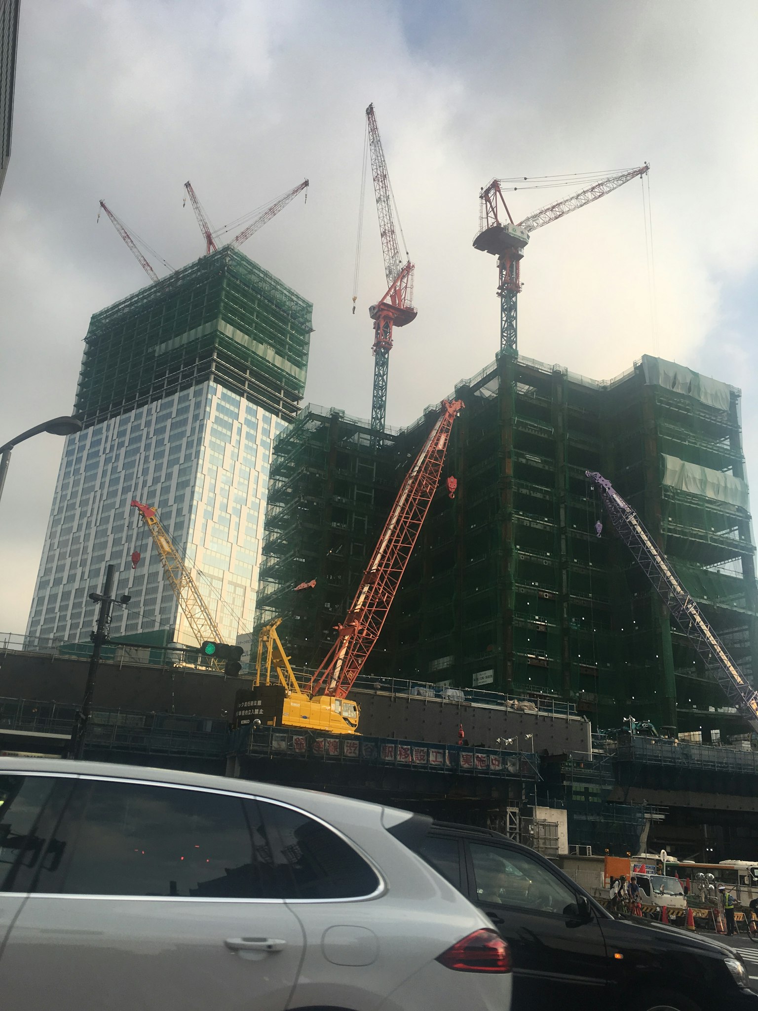 Sitio de construcción con edificios altos y grúas en primer plano un coche es visible con una red verde en las estructuras