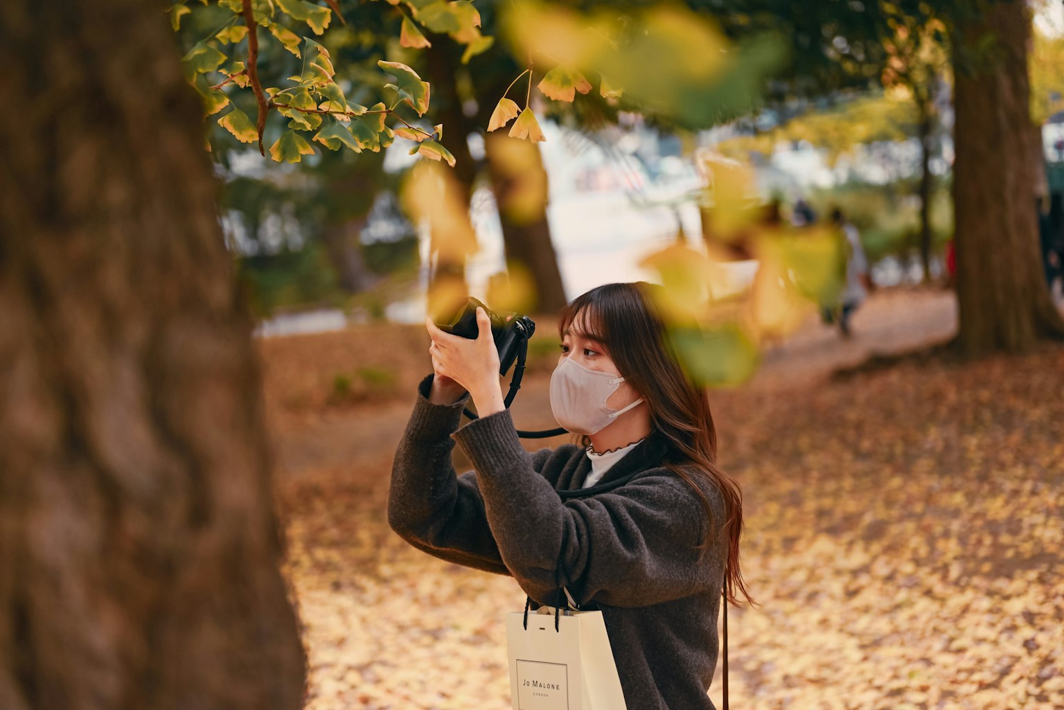 Wanita memegang kamera di taman musim gugur mengenakan masker dikelilingi oleh pohon dan daun yang jatuh