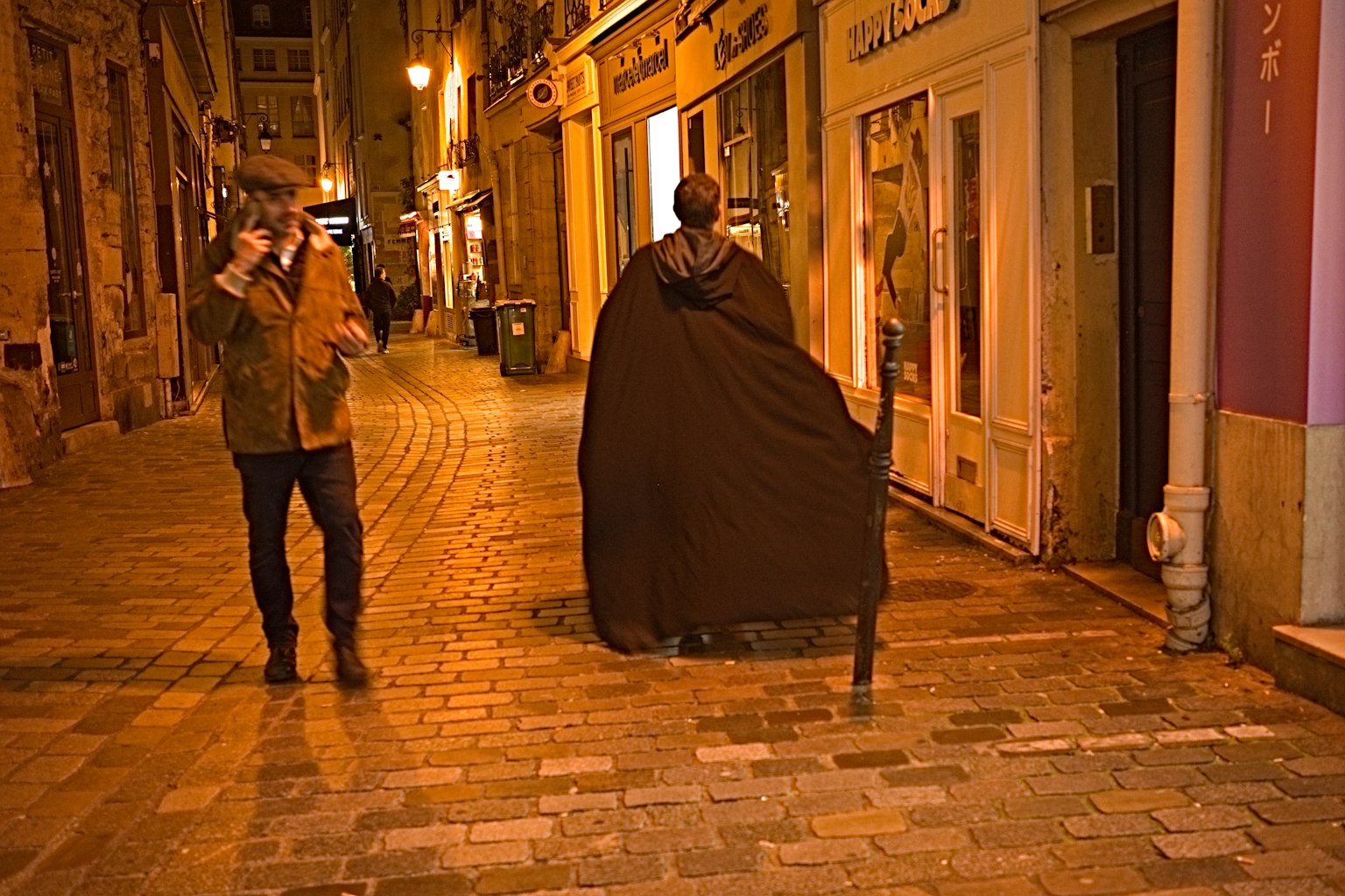 Personne en manteau marchant dans une rue nocturne avec une autre personne tenant un appareil photo