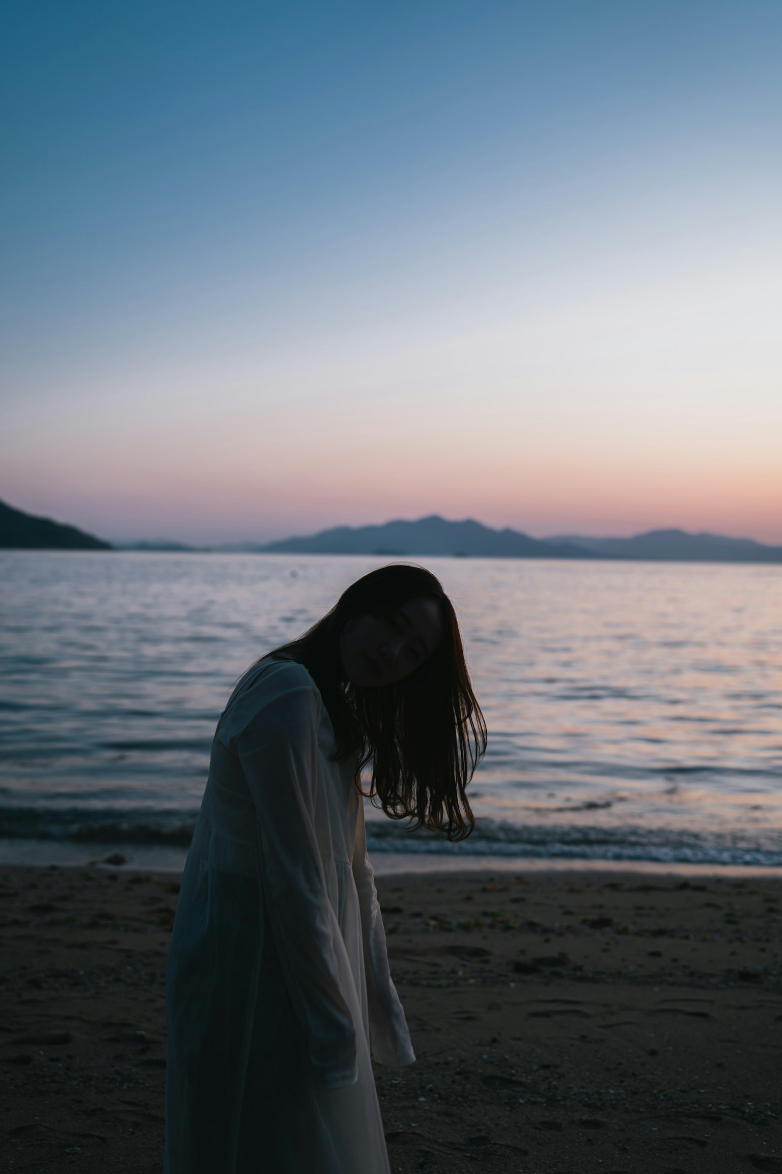 Silhouette einer Frau mit langen Haaren am Strand bei Sonnenuntergang mit ruhigem Meer und Bergen