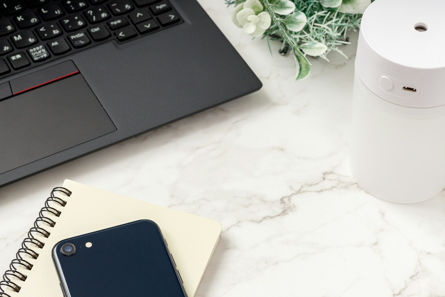 A simple workspace featuring a laptop, white aroma diffuser, smartphone, and notebook on a marble desk