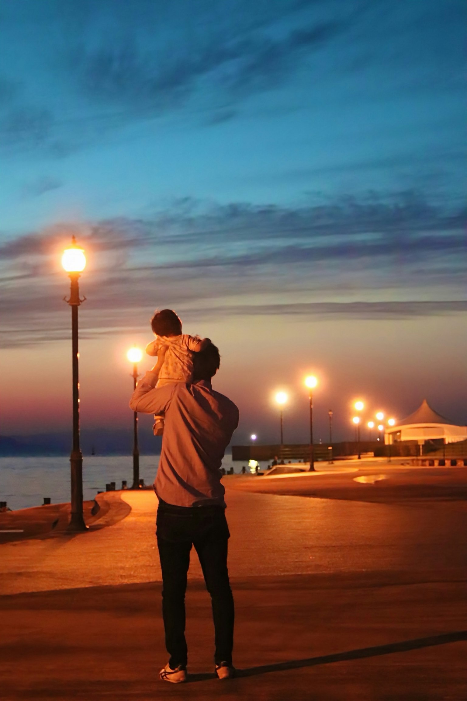 Silhouette eines Vaters, der ein Kind in der Dämmerung am Meer mit Straßenlaternen hält