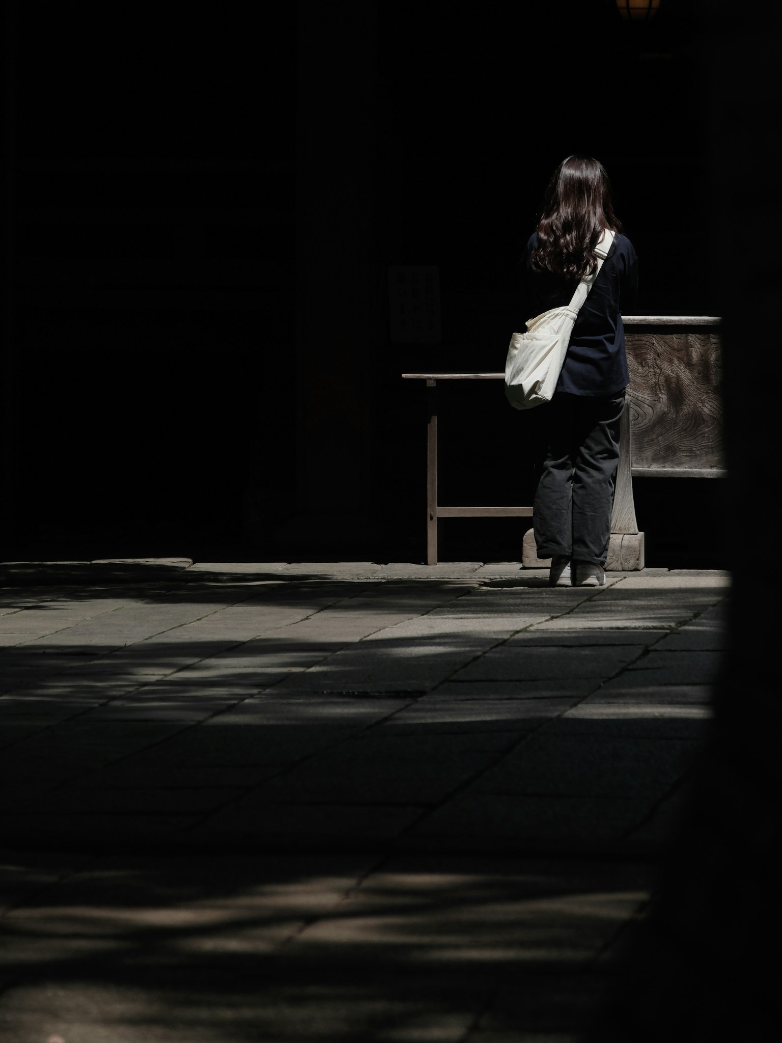 一位女性在陰影中走，背影穿著黑色衣服，背著白色包