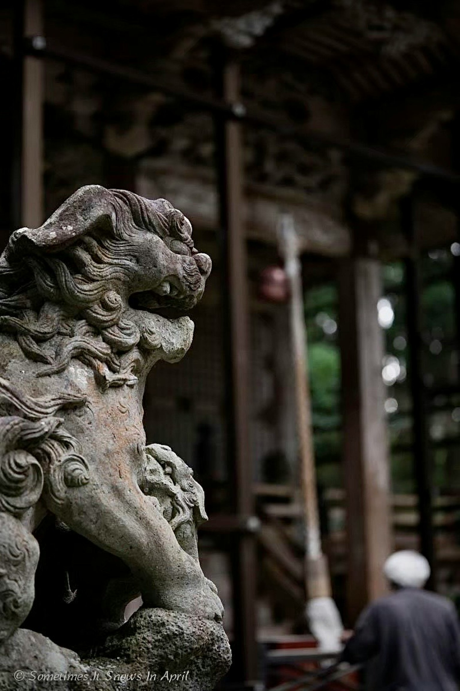 A stone guardian lion statue at a shrine with a blurred figure of a person in the background