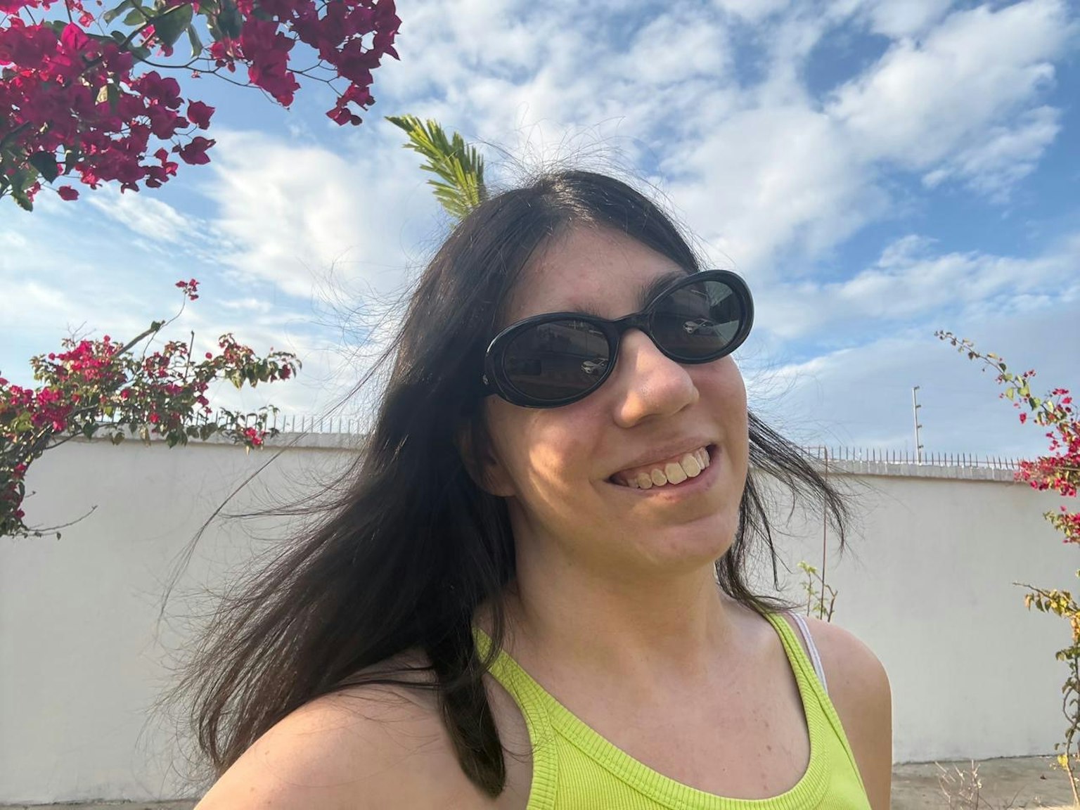 明るい緑のトップを着た女性がサングラスをかけて微笑んでいる背景には青い空と花がある