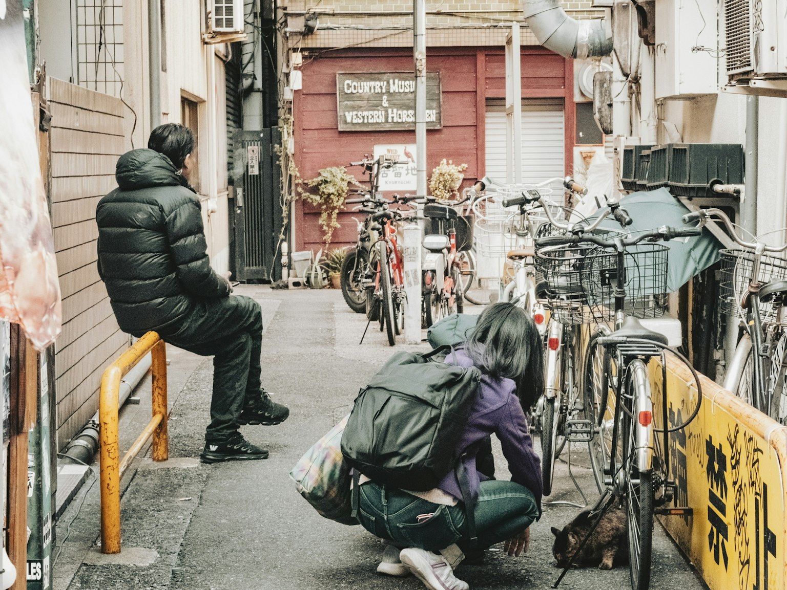 狭窄街道的场景，有一个坐在长椅上的男人和一个蹲在自行车旁边的女人