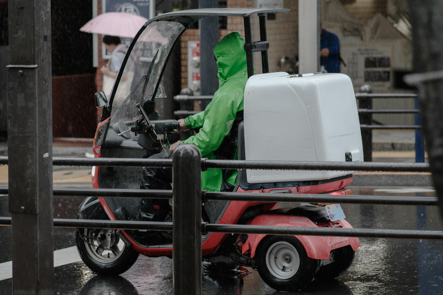 Lieferant in grünem Regenmantel fährt auf einem roten Roller mit weißem Lieferkasten im Regen