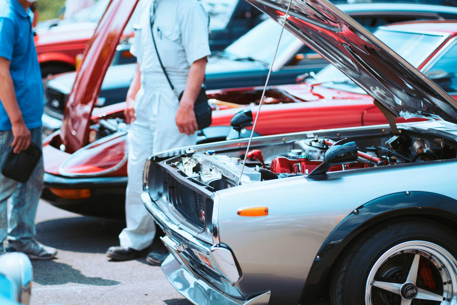 Silver car with open hood showcasing engine surrounded by classic cars and people