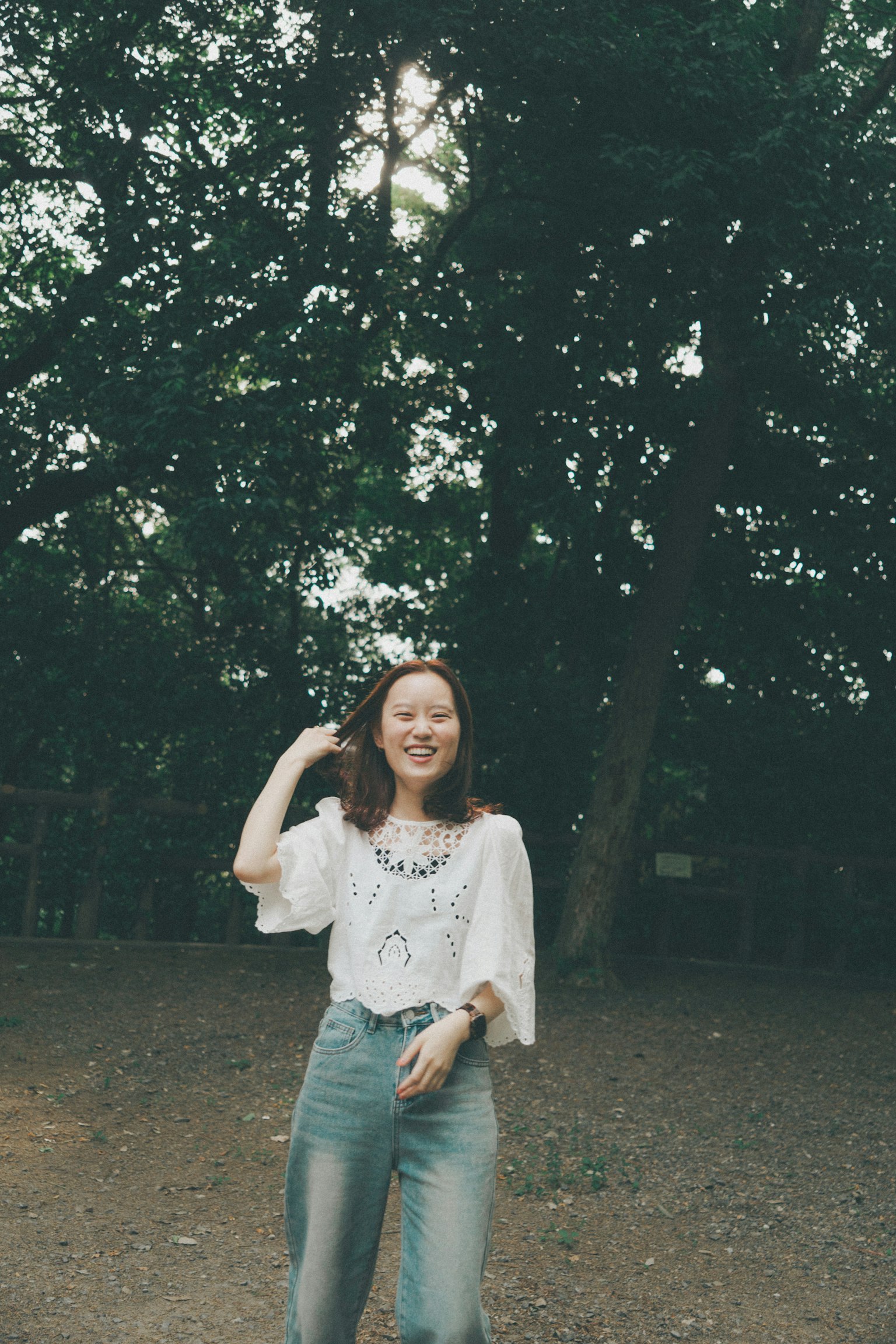Wanita tersenyum berdiri di depan pohon hijau mengenakan atasan putih dan celana jeans